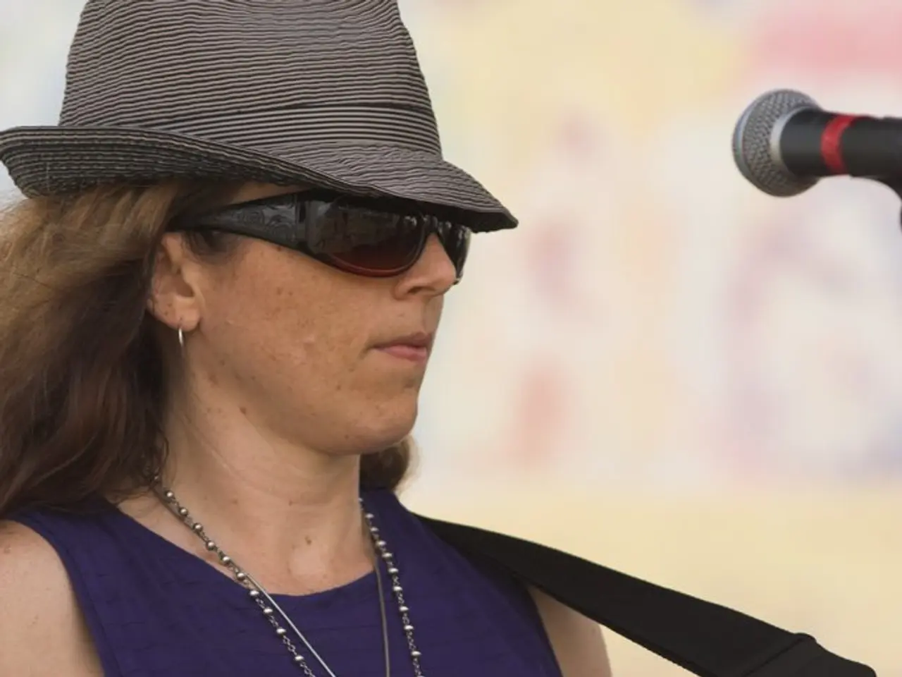 This woman wore purple t-shirt, chain, hat, goggles and playing guitar in-front of mic.