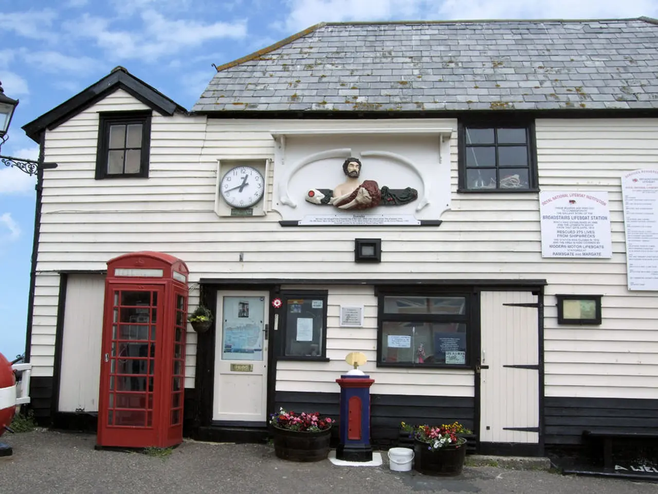 In this image I can see a white colour building, windows, a clock, few boards, a red colour booth,...