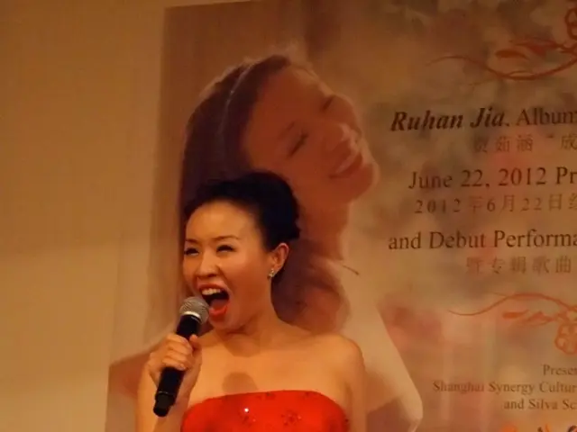 Here we can see a woman standing and singing with a microphone in her hand and behind her we can...